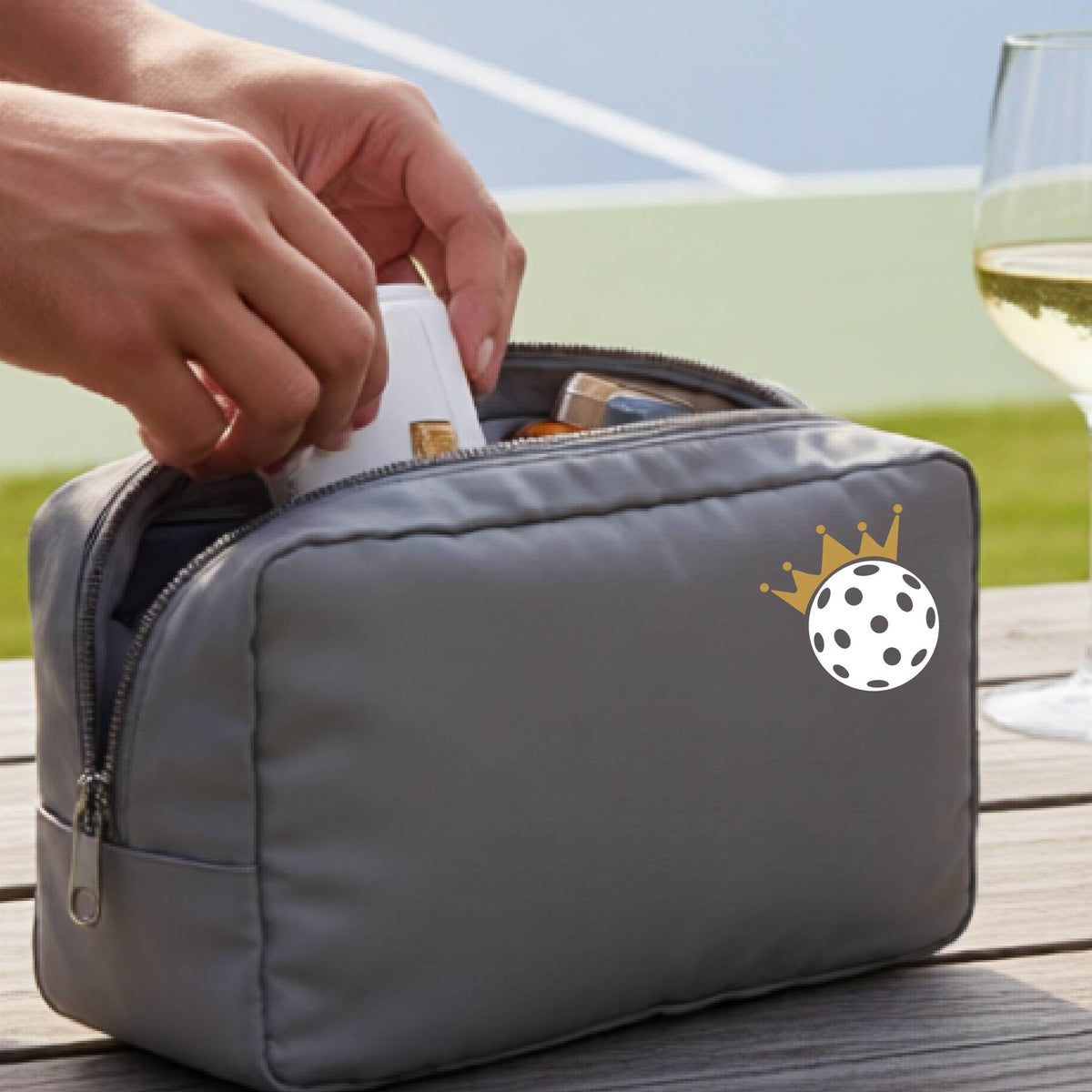 A person holds a gray zippered pouch with a small white pickleball and gold crown graphic on the front, set against an outdoor background with a pickleball court and wooden table.