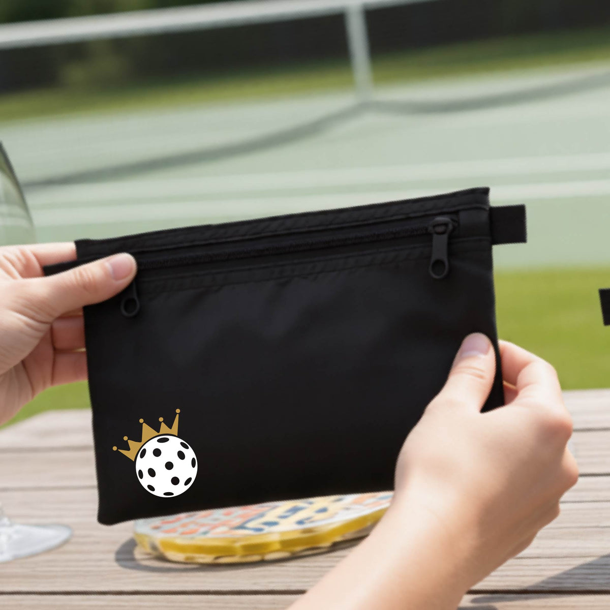 A person holds a black zippered pouch with a small white pickleball and gold crown graphic on the front, set against an outdoor background with a pickleball court and wooden table.