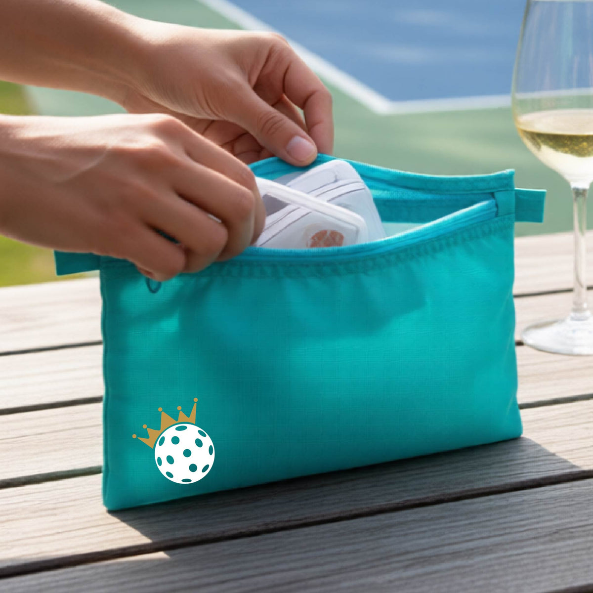 A person holds a turquoise zippered pouch with a small white pickleball and gold crown graphic on the front, set against an outdoor background with a pickleball court and wooden table.