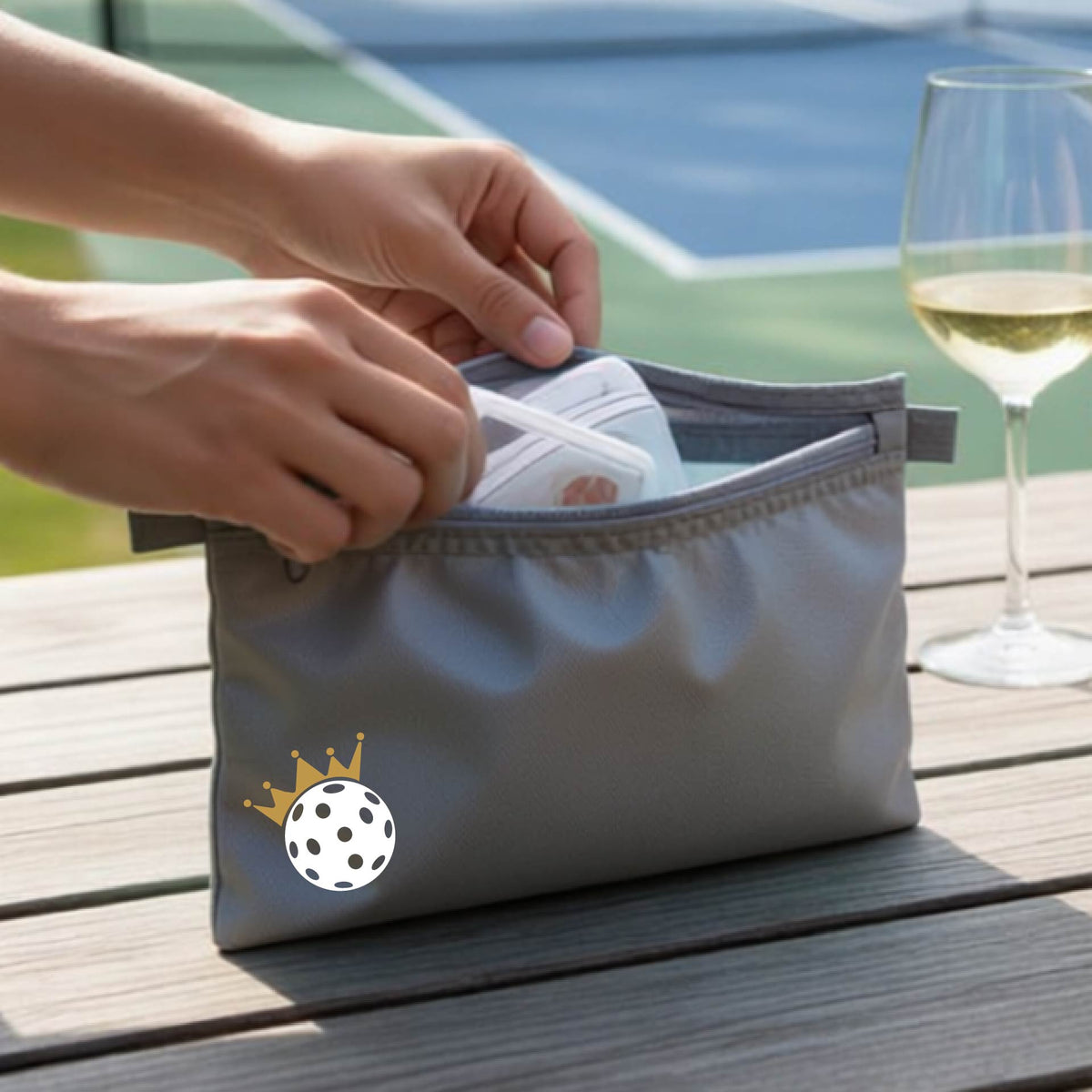 A person holds a gray zippered pouch with a small white pickleball and gold crown graphic on the front, set against an outdoor background with a pickleball court and wooden table.