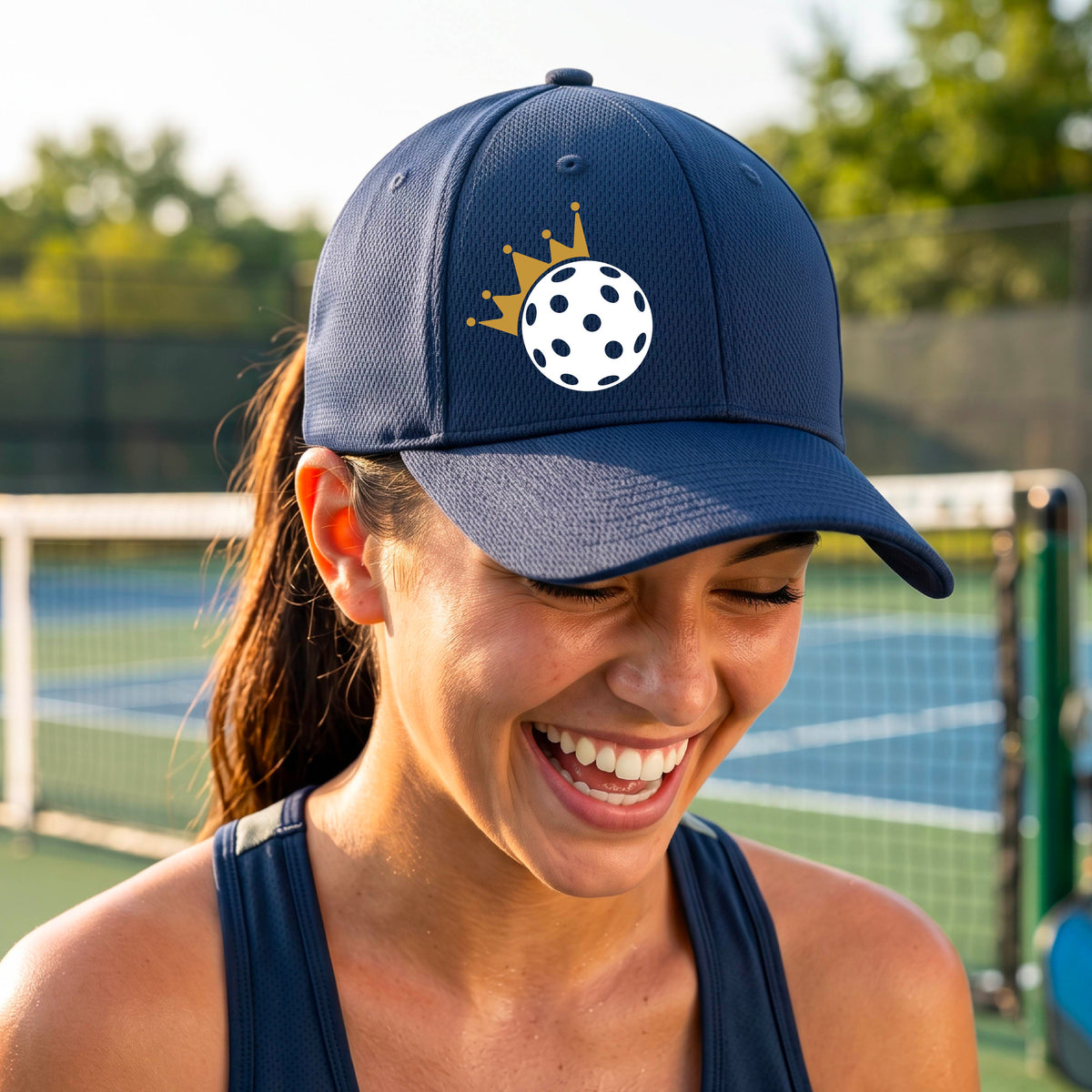 A person outdoors wearing a navy blue baseball cap with a white pickleball graphic of small gold crown on the front.