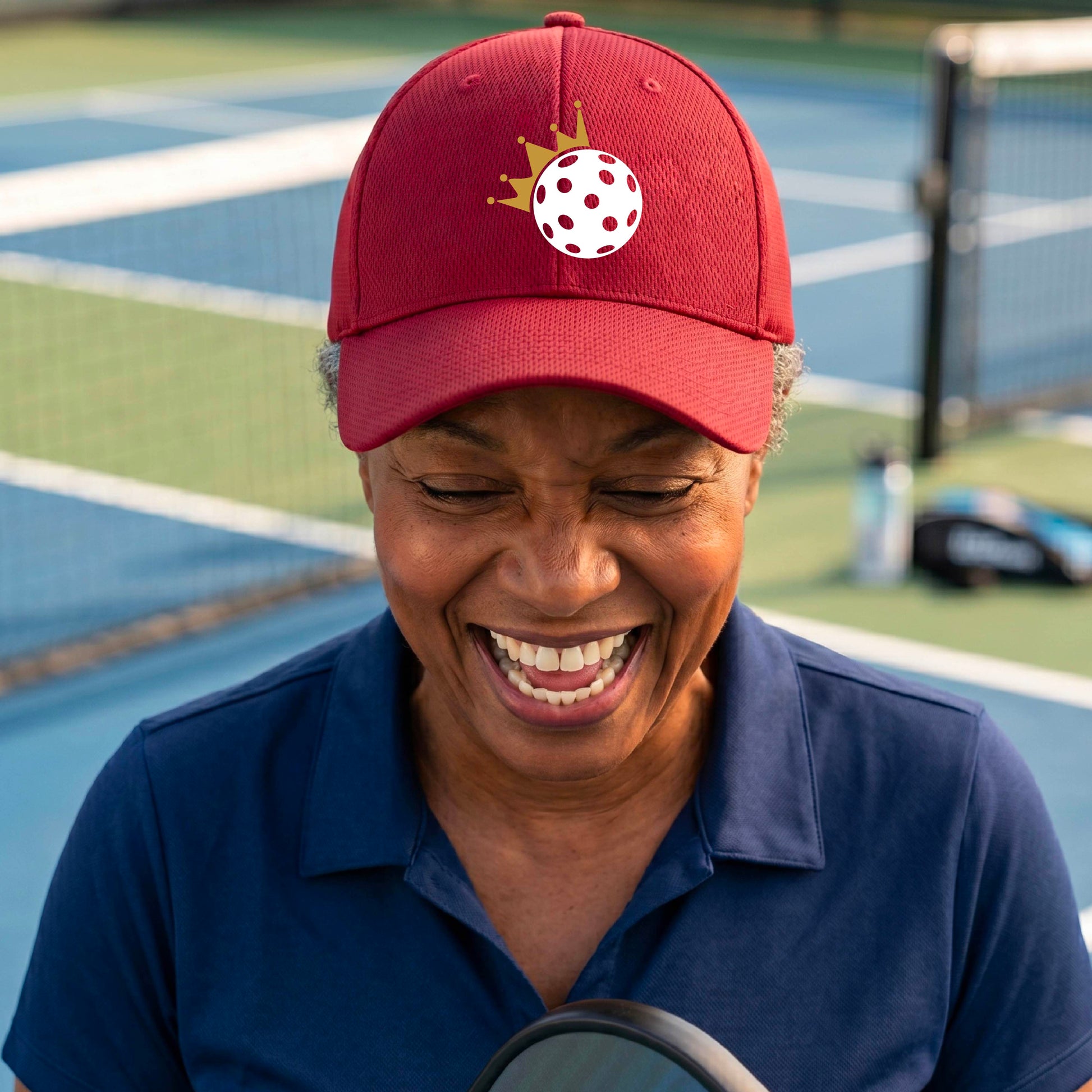 A person outdoors wearing a red baseball cap with a white pickleball graphic of small gold crown on the front.