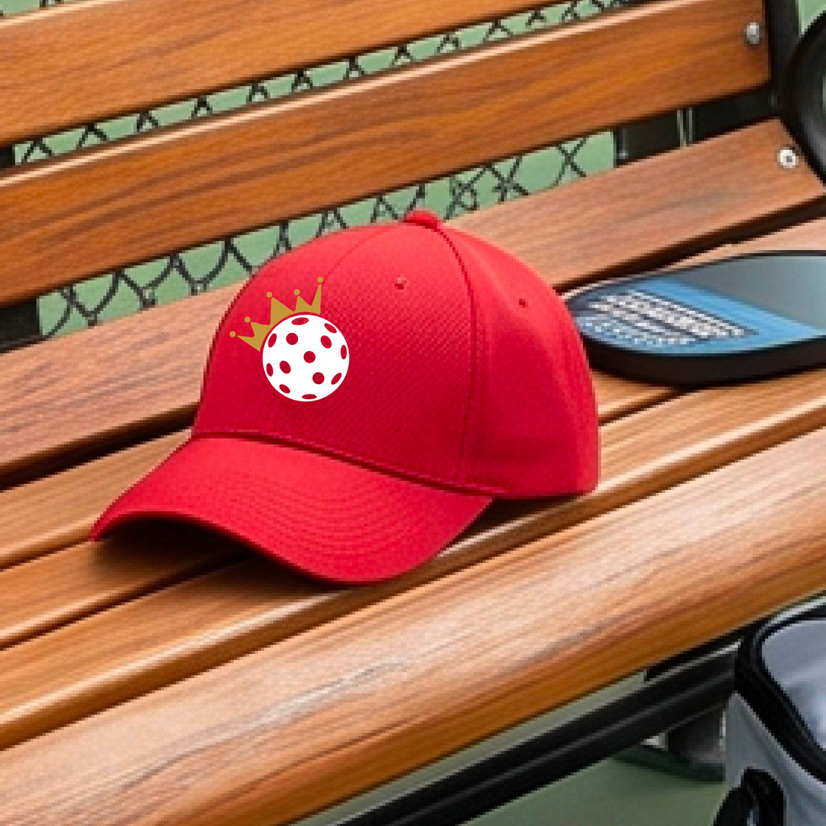 A person outdoors wearing a red baseball cap with a white pickleball graphic of small gold crown on the front.