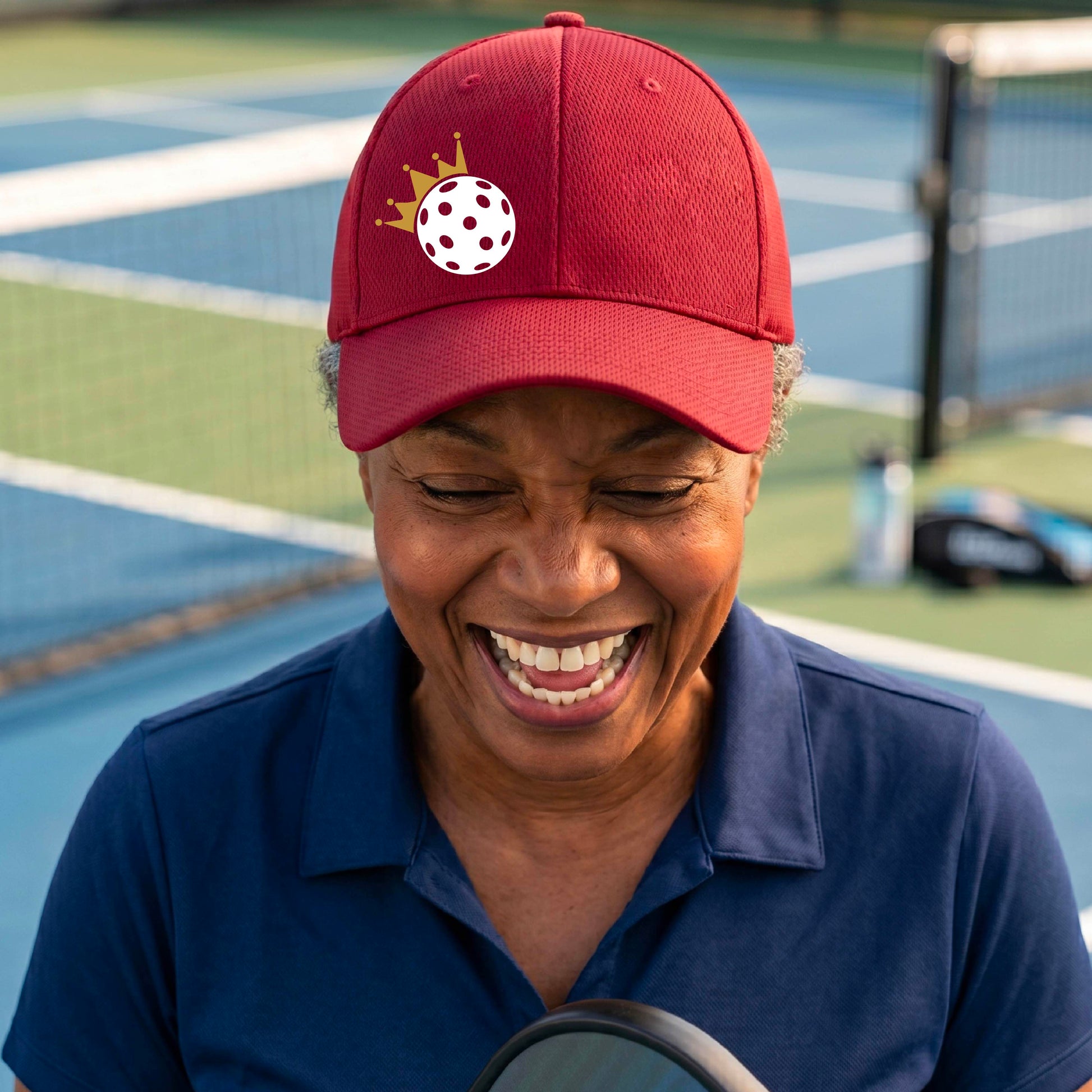 A person outdoors wearing a red baseball cap with a white pickleball graphic of small gold crown on the front.