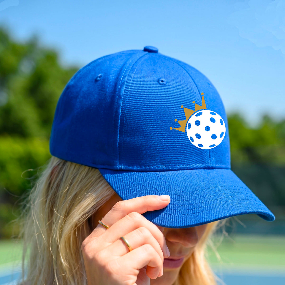 A person outdoors wearing a bright blue baseball cap with a white pickleball graphic of small gold crown on the front.