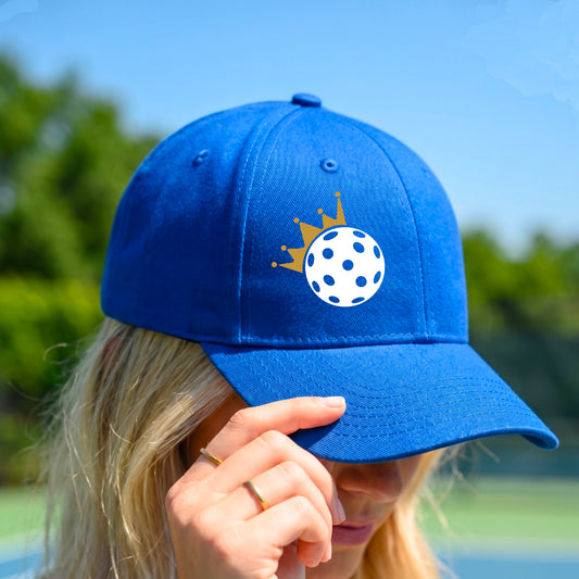 A person outdoors wearing a bright blue baseball cap with a white pickleball graphic of small gold crown on the front.