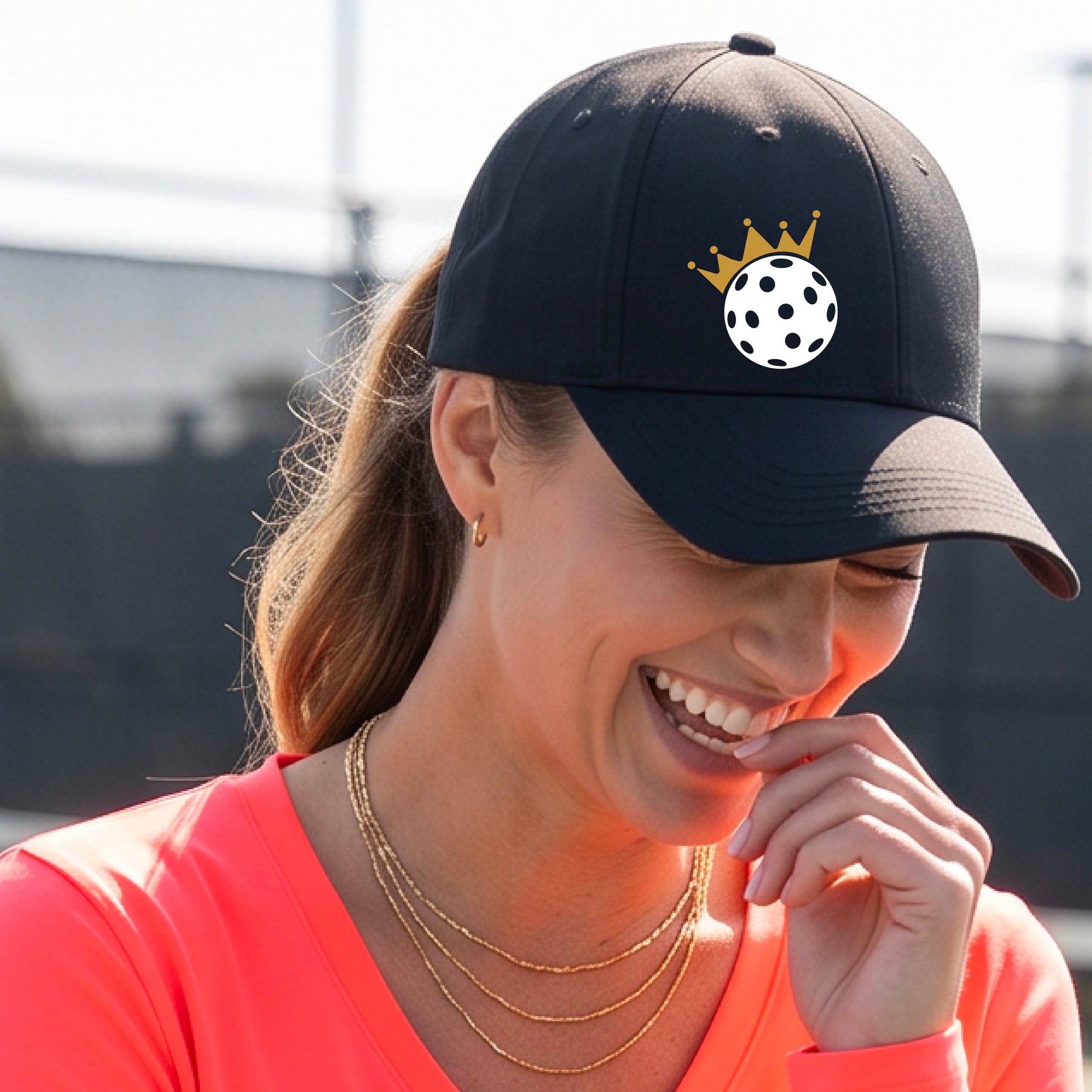 A person outdoors wearing a black baseball cap with a white pickleball graphic of small gold crown on the front.