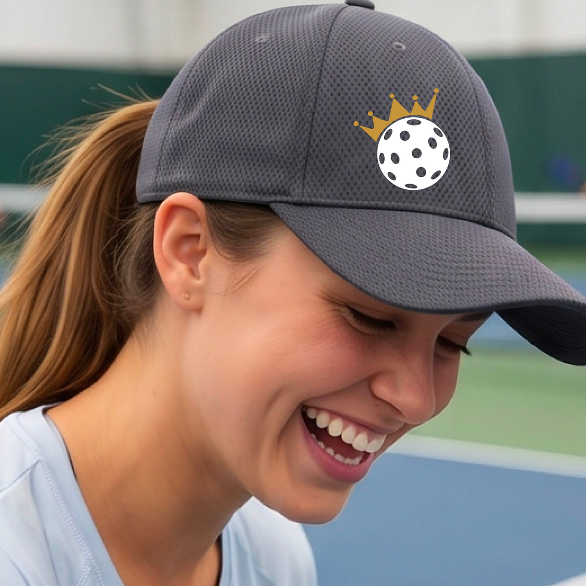 A person outdoors wearing a gray baseball cap with a white pickleball graphic of small gold crown on the front.