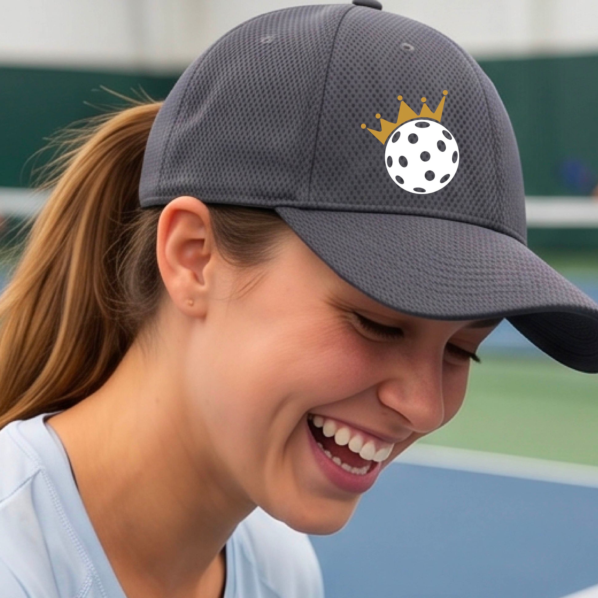 A person outdoors wearing a gray baseball cap with a white pickleball graphic of small gold crown on the front.