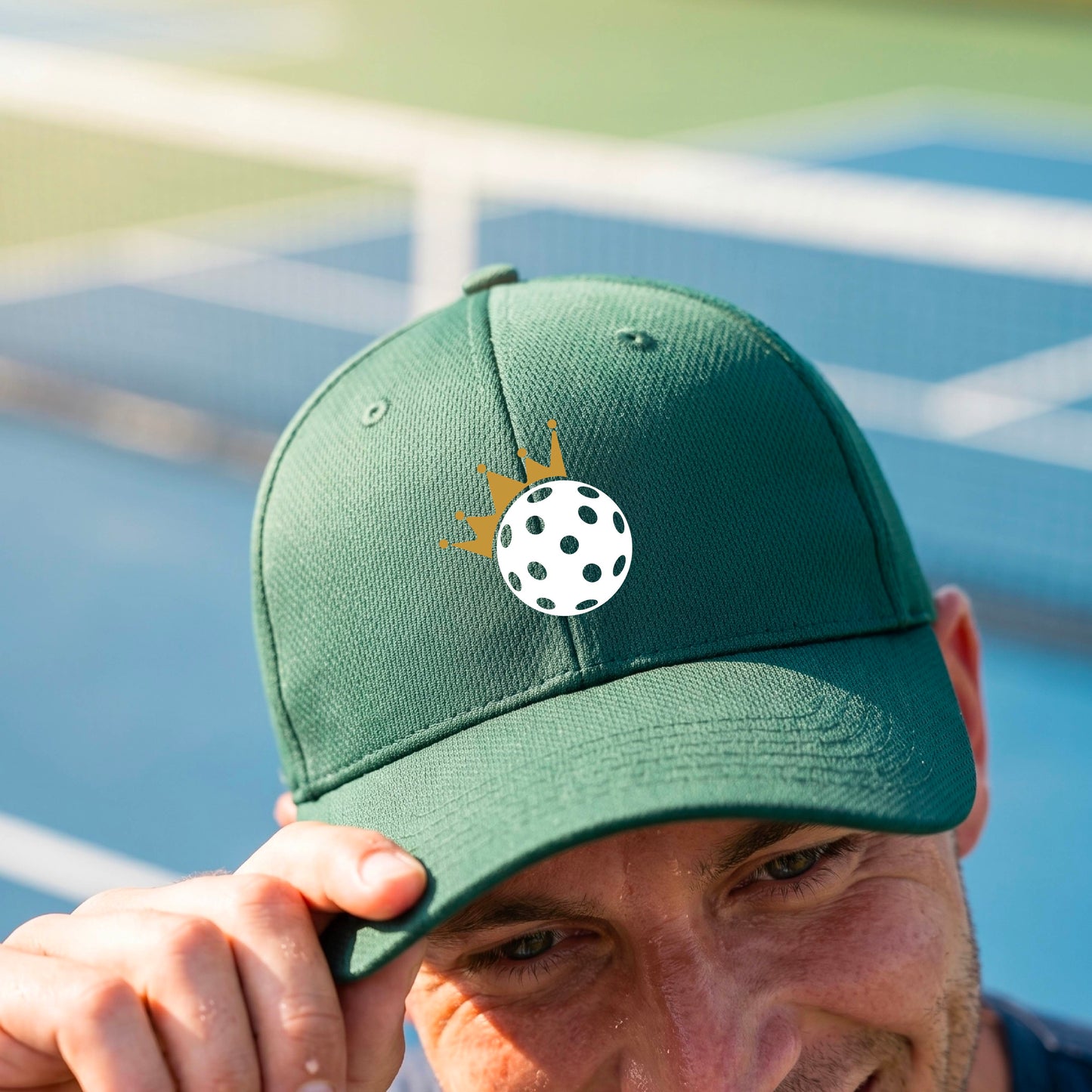A person outdoors wearing a green baseball cap with a white pickleball graphic of small gold crown on the front.