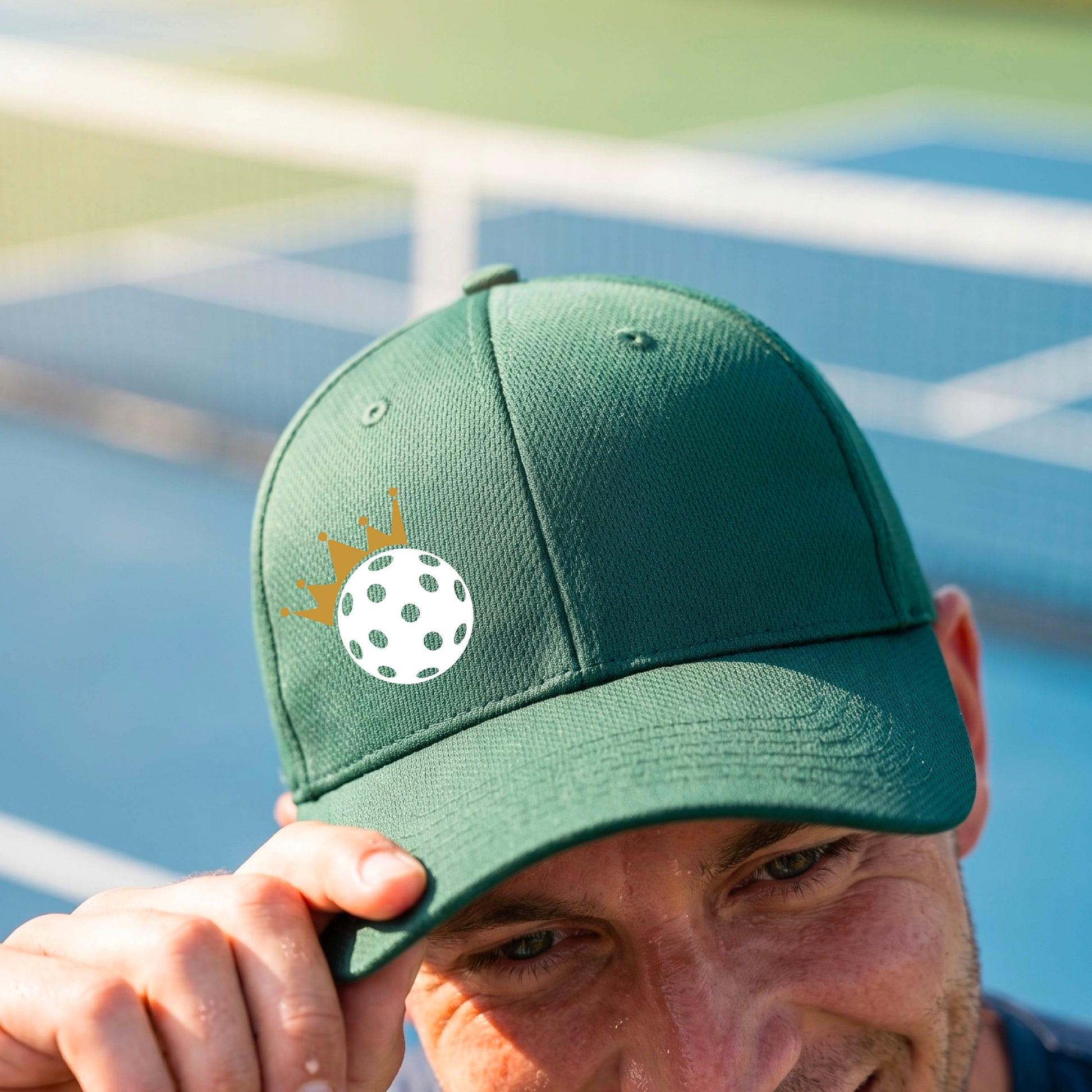 A person outdoors wearing a green baseball cap with a white pickleball graphic of small gold crown on the front.