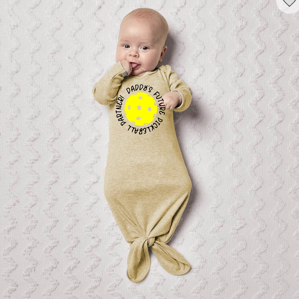 A baby sleeping on a white blanket on a pickleball court, with a graphic text of  'Daddy's Future Pickleball Partner" wearing a beige footed knotted newborn sleeper.