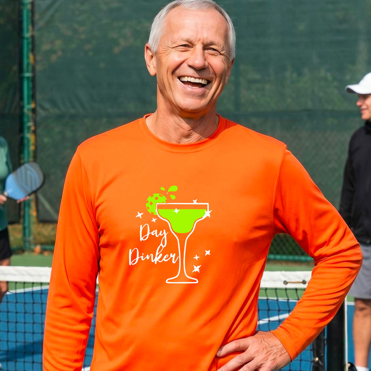 A smiling man on an outdoor pickleball court wearing a bright orange long-sleeve shirt with a 'Day Dinker' martini graphic with a pickleball lime garnish. 