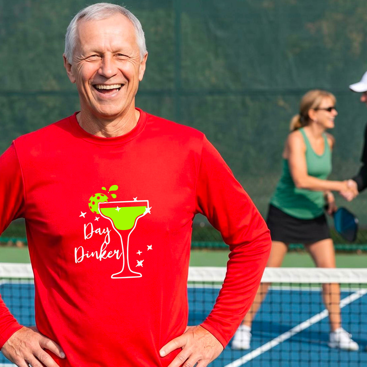 A smiling man on an outdoor pickleball court wearing a bright red long-sleeve shirt with a 'Day Dinker' martini graphic with a pickleball lime garnish. 