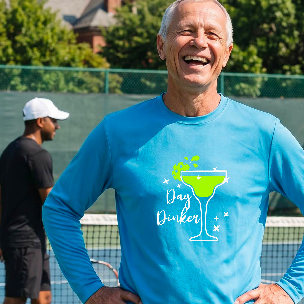A smiling man on an outdoor pickleball court wearing a bright blue long-sleeve shirt with a 'Day Dinker' martini graphic with a pickleball lime garnish. 