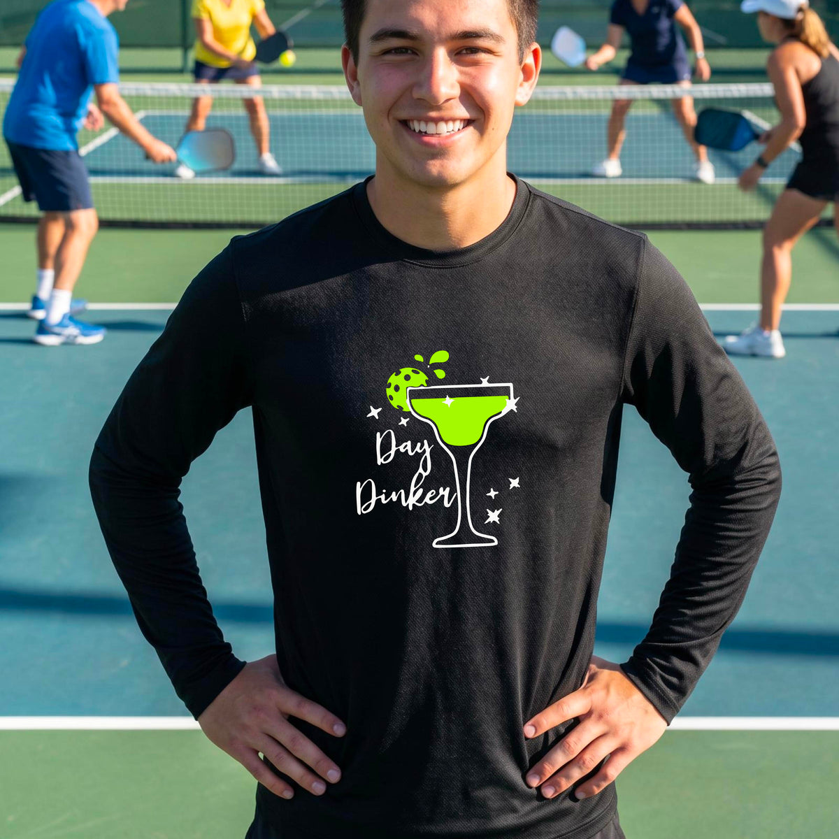 A smiling man on an outdoor pickleball court wearing a black long-sleeve shirt with a 'Day Dinker' martini graphic with a pickleball lime garnish. 