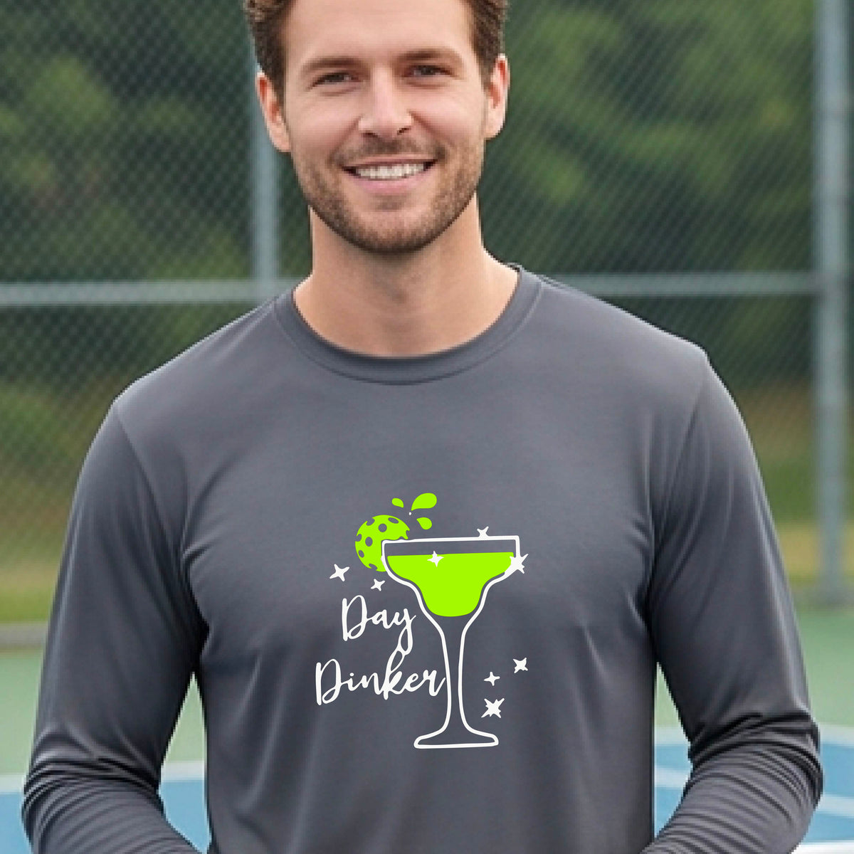 A smiling man on an outdoor pickleball court wearing a gray long-sleeve shirt with a 'Day Dinker' martini graphic with a pickleball lime garnish. 