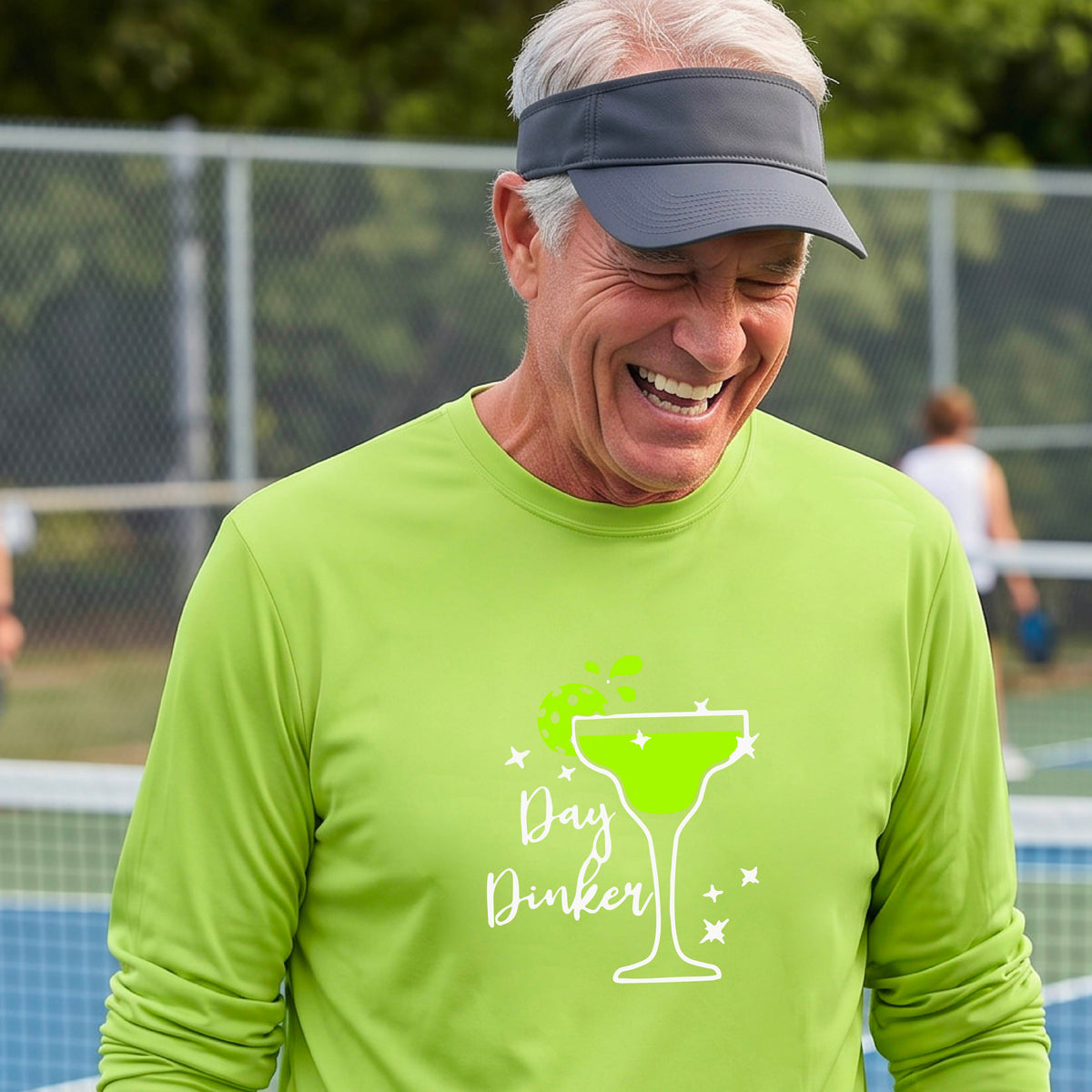 A smiling man on an outdoor pickleball court wearing a bright lime green long-sleeve shirt with a 'Day Dinker' martini graphic with a pickleball lime garnish. 