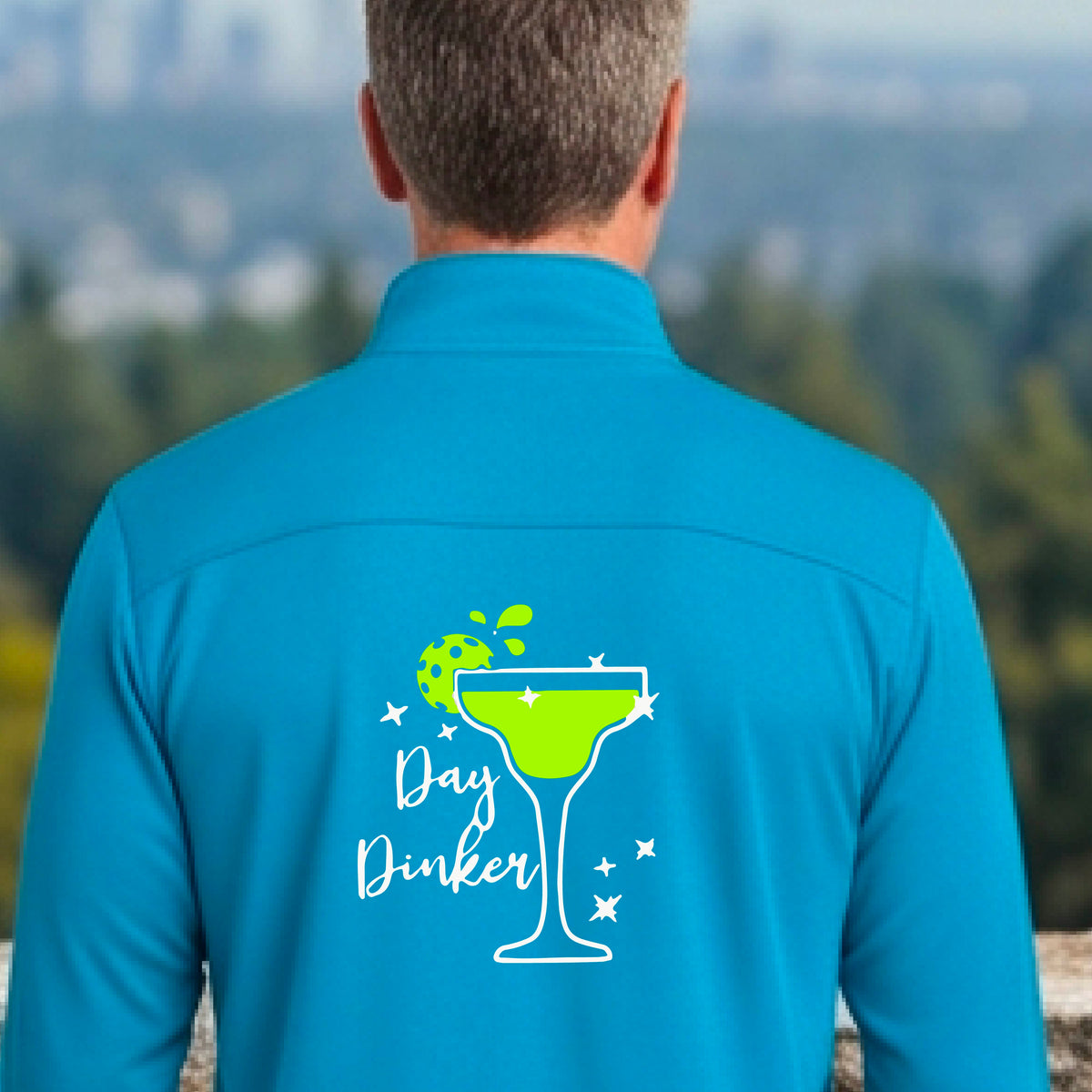 A man viewed from behind wearing a bright blue quarter-zip pullover with a white and green 'Day Dinker' cocktail graphic with a green pickleball lime garnish, on the back, standing on a pickleball court.