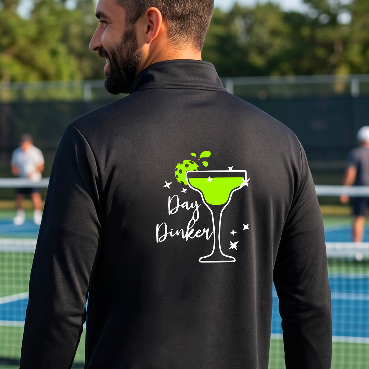 A man viewed from behind wearing a black quarter-zip pullover with a white and green 'Day Dinker' cocktail graphic with a green pickleball lime garnish, on the back, standing on a pickleball court.