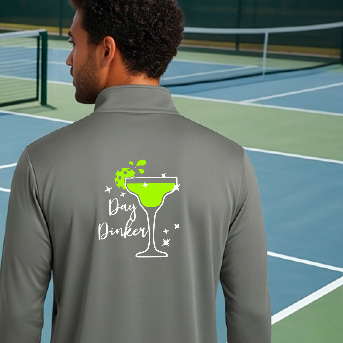 A man viewed from behind wearing a gray quarter-zip pullover with a white and green 'Day Dinker' cocktail graphic with a green pickleball lime garnish, on the back, standing on a pickleball court.
