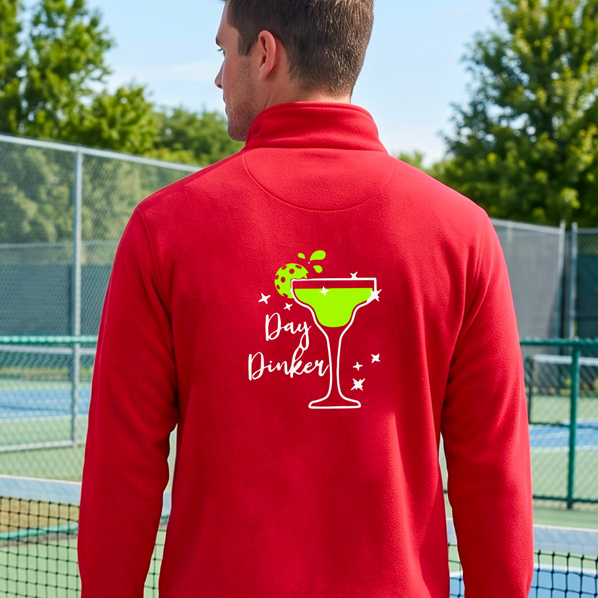 A man viewed from behind wearing a bright red quarter-zip pullover with a white and green 'Day Dinker' cocktail graphic with a green pickleball lime garnish, on the back, standing on a pickleball court.