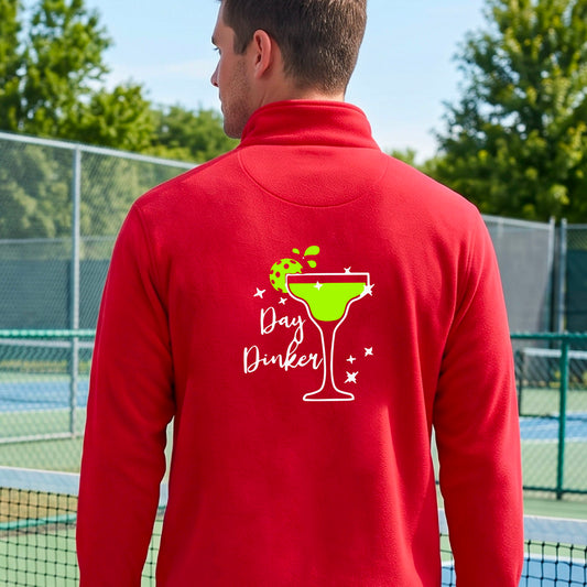 A man viewed from behind wearing a bright red quarter-zip pullover with a white and green 'Day Dinker' cocktail graphic with a green pickleball lime garnish, on the back, standing on a pickleball court.