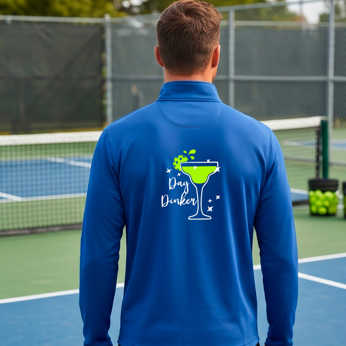 A man viewed from behind wearing a bright royal blue quarter-zip pullover with a white and green 'Day Dinker' cocktail graphic with a green pickleball lime garnish, on the back, standing on a pickleball court.