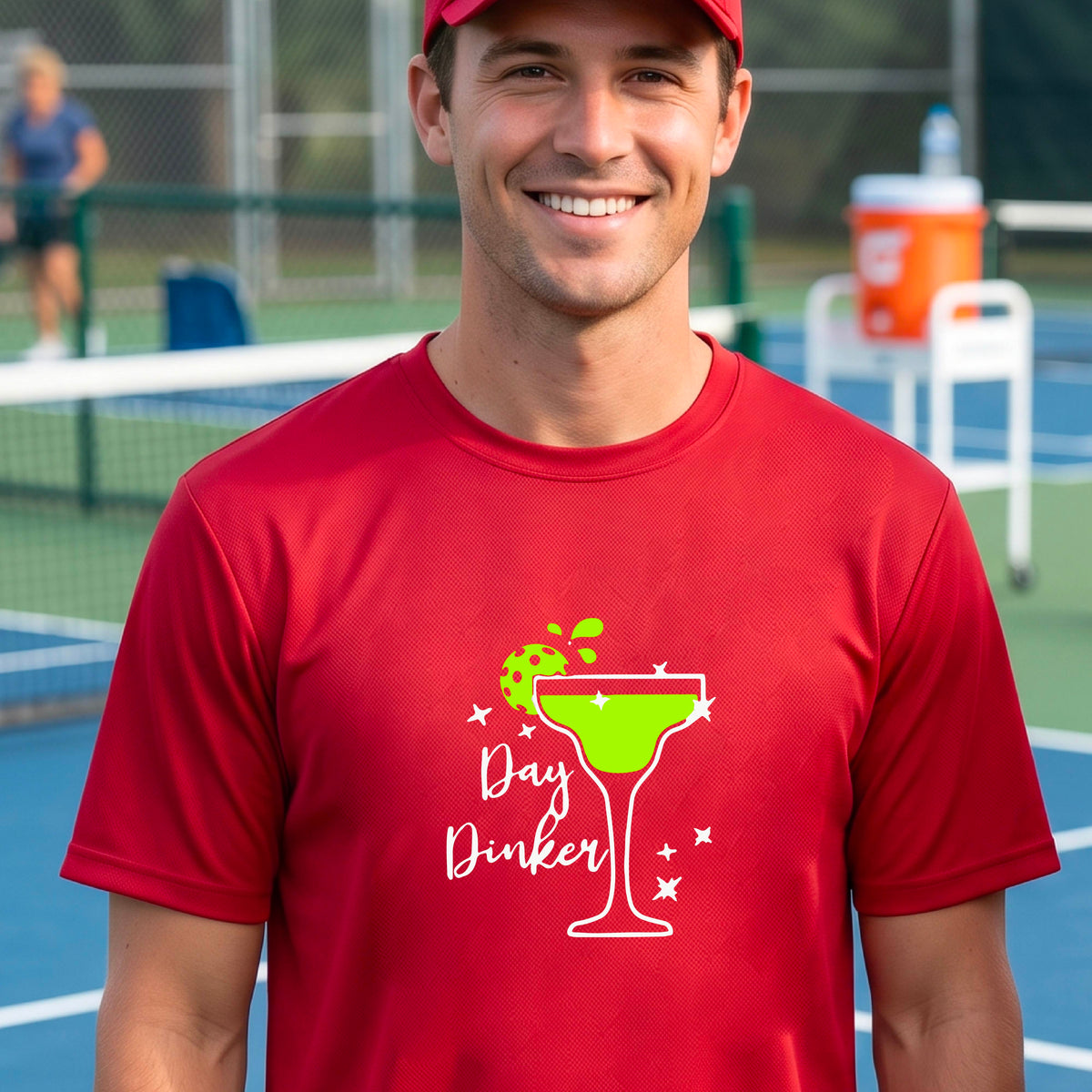 A man laughing on a pickleball court wearing a red t-shirt with a 'Day Dinker' martini with a pickleball lime garnish graphic in green and white.