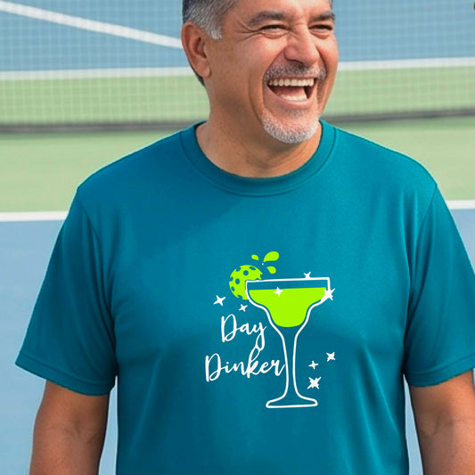 A man laughing on a pickleball court wearing a teal t-shirt with a 'Day Dinker' martini with a pickleball lime garnish graphic in green and white.
