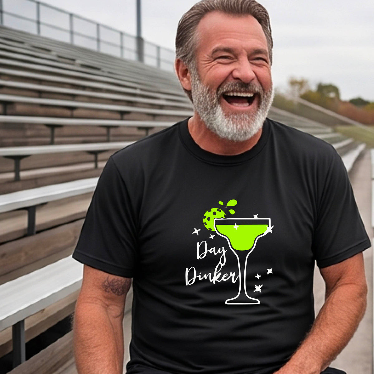A smiling man wearing a black graphic t-shirt with a green and white 'Day Dinker' cocktail design with a green pickleball lime garnish. 