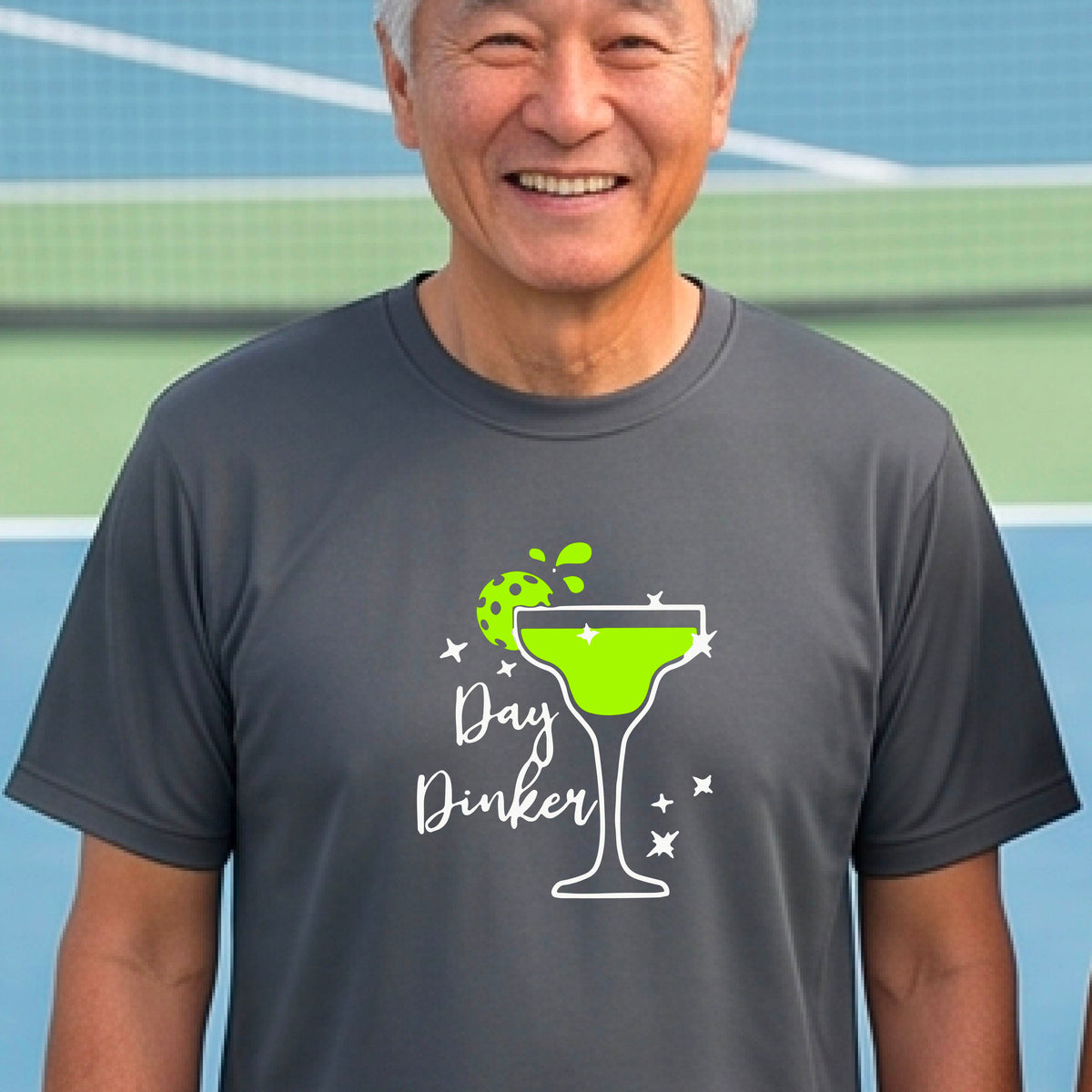 A man laughing on a pickleball court wearing a gray t-shirt with a 'Day Dinker' martini with a pickleball lime garnish graphic in green and white.