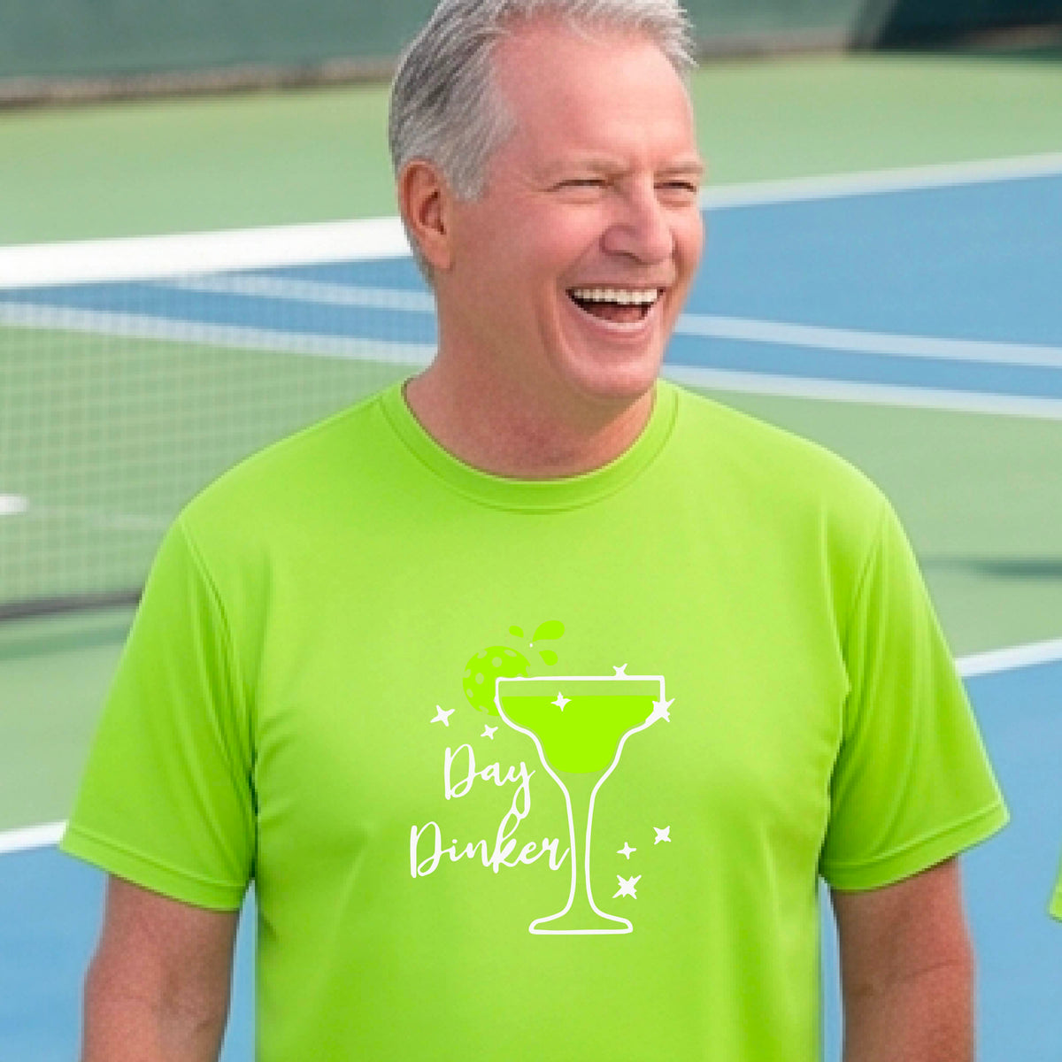 A man laughing on a pickleball court wearing a lime green t-shirt with a 'Day Dinker' martini with a pickleball lime garnish graphic in green and white.