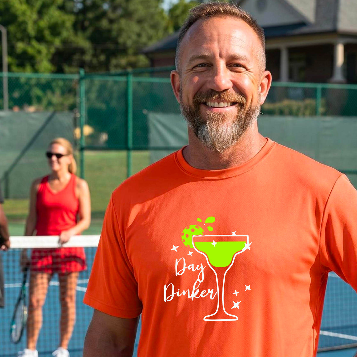A man laughing on a pickleball court wearing an orange t-shirt with a 'Day Dinker' martini with a pickleball lime garnish graphic in green and white.