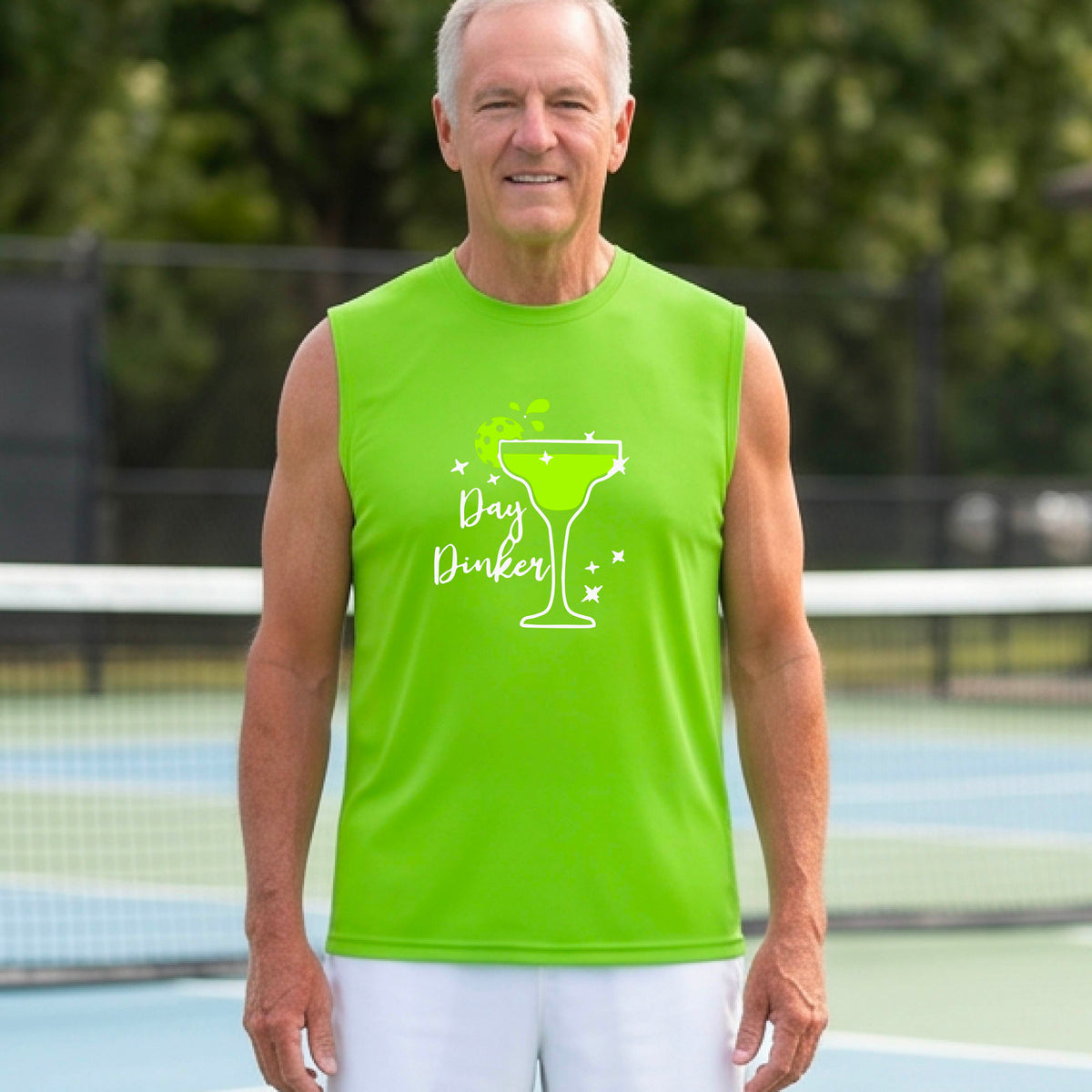 A man smiling on an outdoor pickleball court wearing a lime green sleeveless tank top with a green cocktail martini glass with a pickleball lim garnish graphic and the words 'Day Dinkster' printed on the front. 