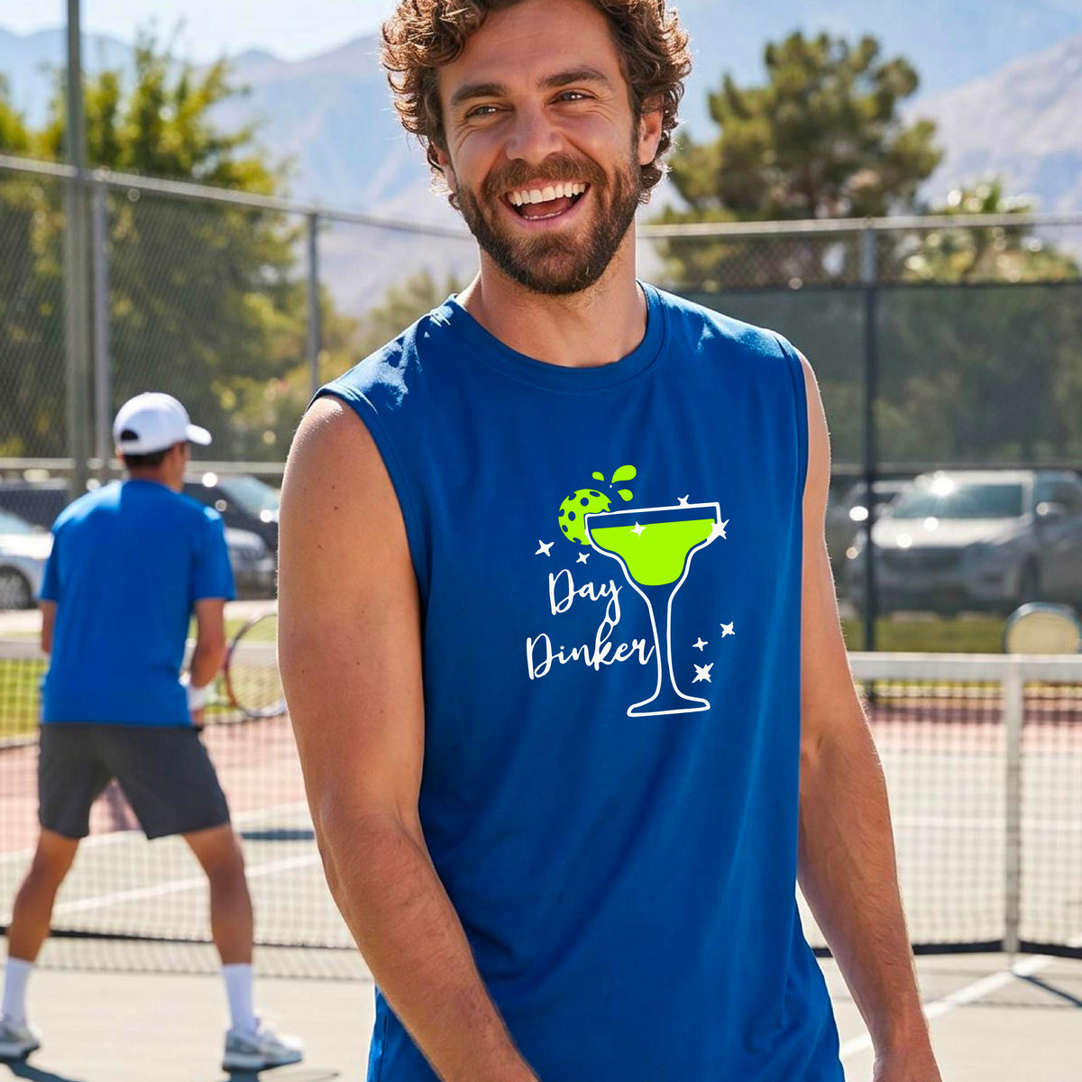 A man smiling on an outdoor pickleball court wearing a royal blue sleeveless tank top with a green cocktail martini glass with a pickleball lim garnish graphic and the words 'Day Dinkster' printed on the front. 