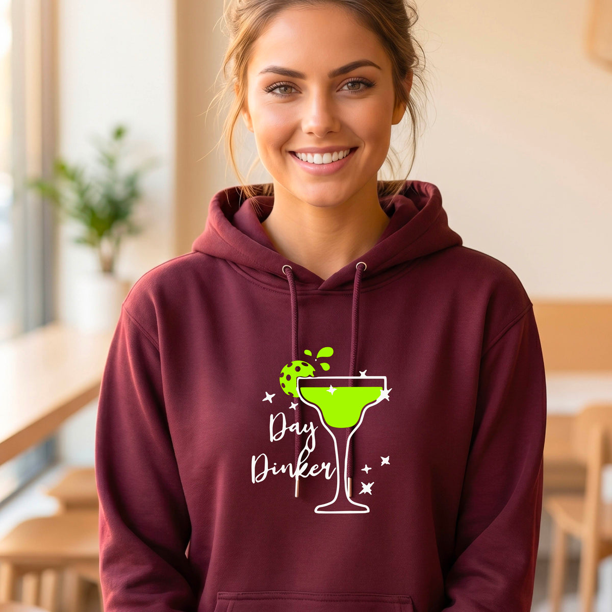 A smiling woman wearing a maroon hoodie with a green margarita glass graphic and green pickleball lime garnish and the words 'Day Dinker' on the front.