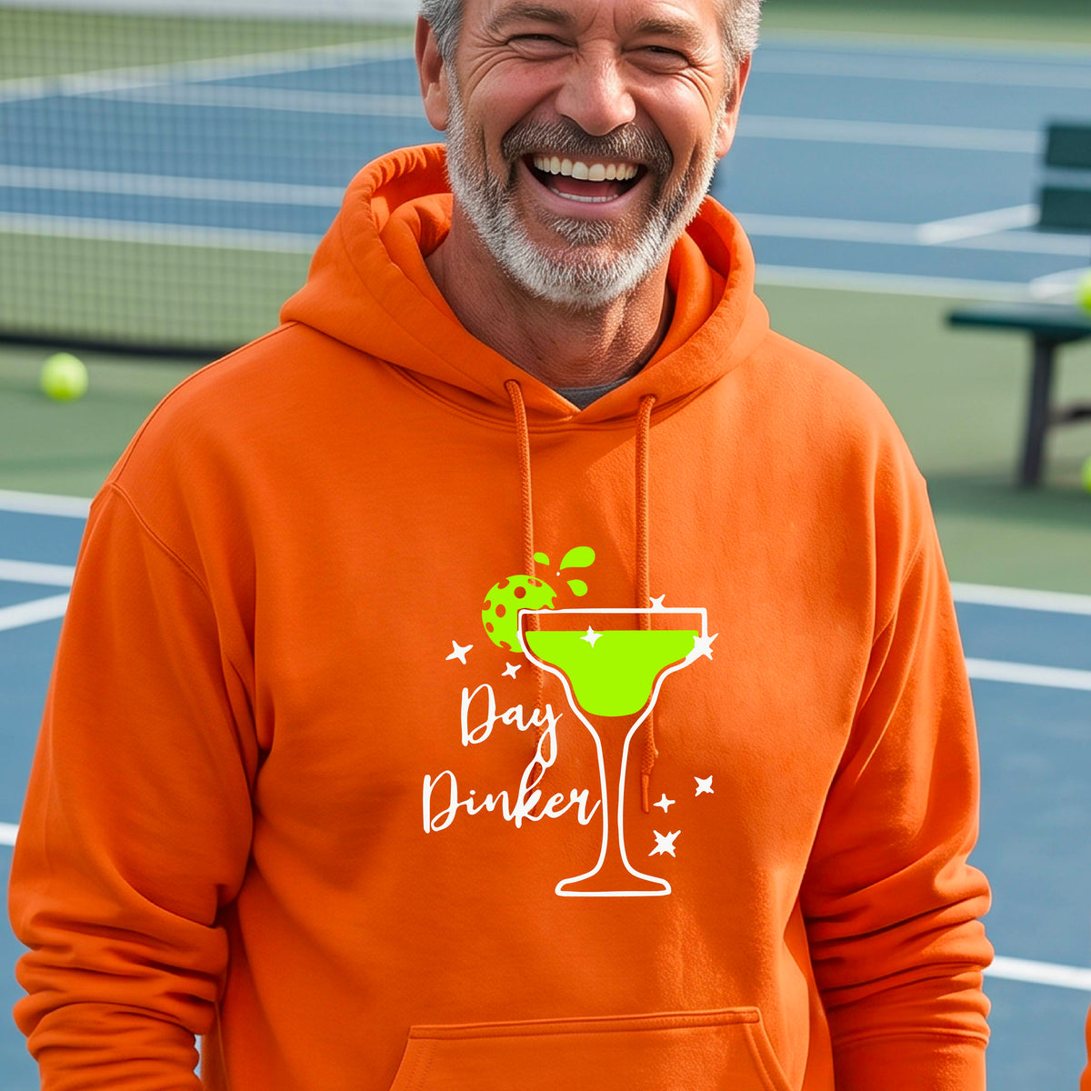 A smiling man wearing an orange hoodie with a green margarita glass graphic and green pickleball lime garnish and the words 'Day Dinker' on the front.