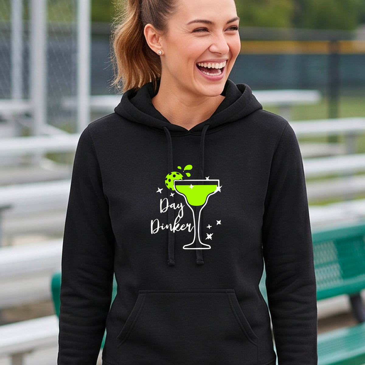 A woman wearing a black hoodie with a 'Day Dinker' martini graphic in green, white, and lime green pickleball lime garish, set against a blurred pickleball court background.