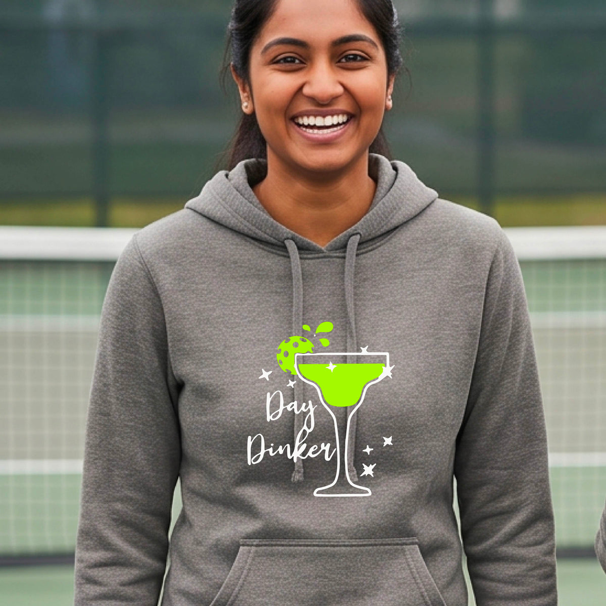 A woman wearing a gray hoodie with a 'Day Dinker' martini graphic in green, white, and lime green pickleball lime garish, set against a blurred pickleball court background.