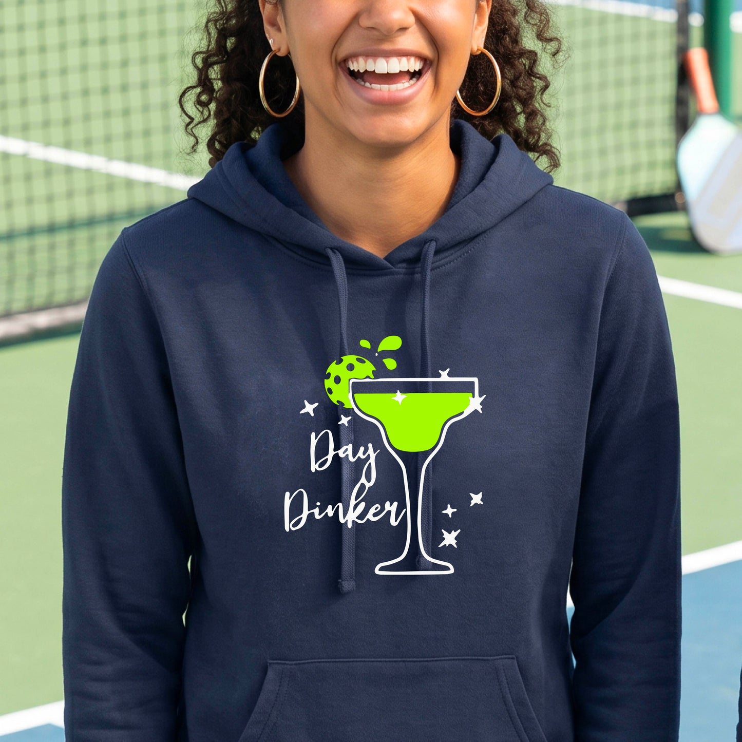 A woman wearing a navy blue hoodie with a 'Day Dinker' martini graphic in green, white, and lime green pickleball lime garish, set against a blurred pickleball court background.