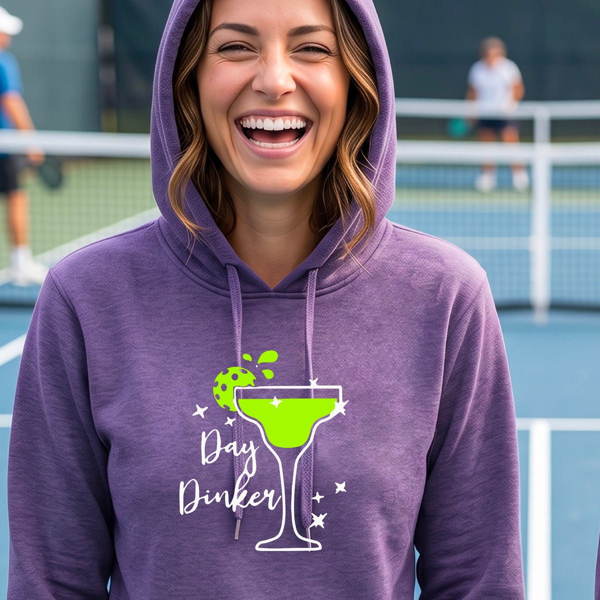 A woman wearing a purple hoodie with a 'Day Dinker' martini graphic in green, white, and lime green pickleball lime garish, set against a blurred pickleball court background.