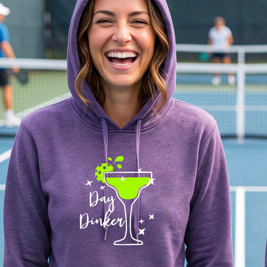 A woman wearing a purple hoodie with a 'Day Dinker' martini graphic in green, white, and lime green pickleball lime garish, set against a blurred pickleball court background.