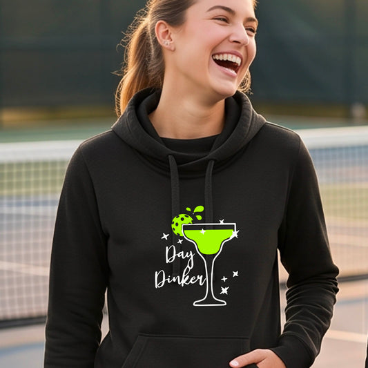 A woman laughing on a pickleball court wearing a black hoodie with a green cocktail glass graphic and green pickleball lime garnish and the words 'Day Dinker' printed on the front.