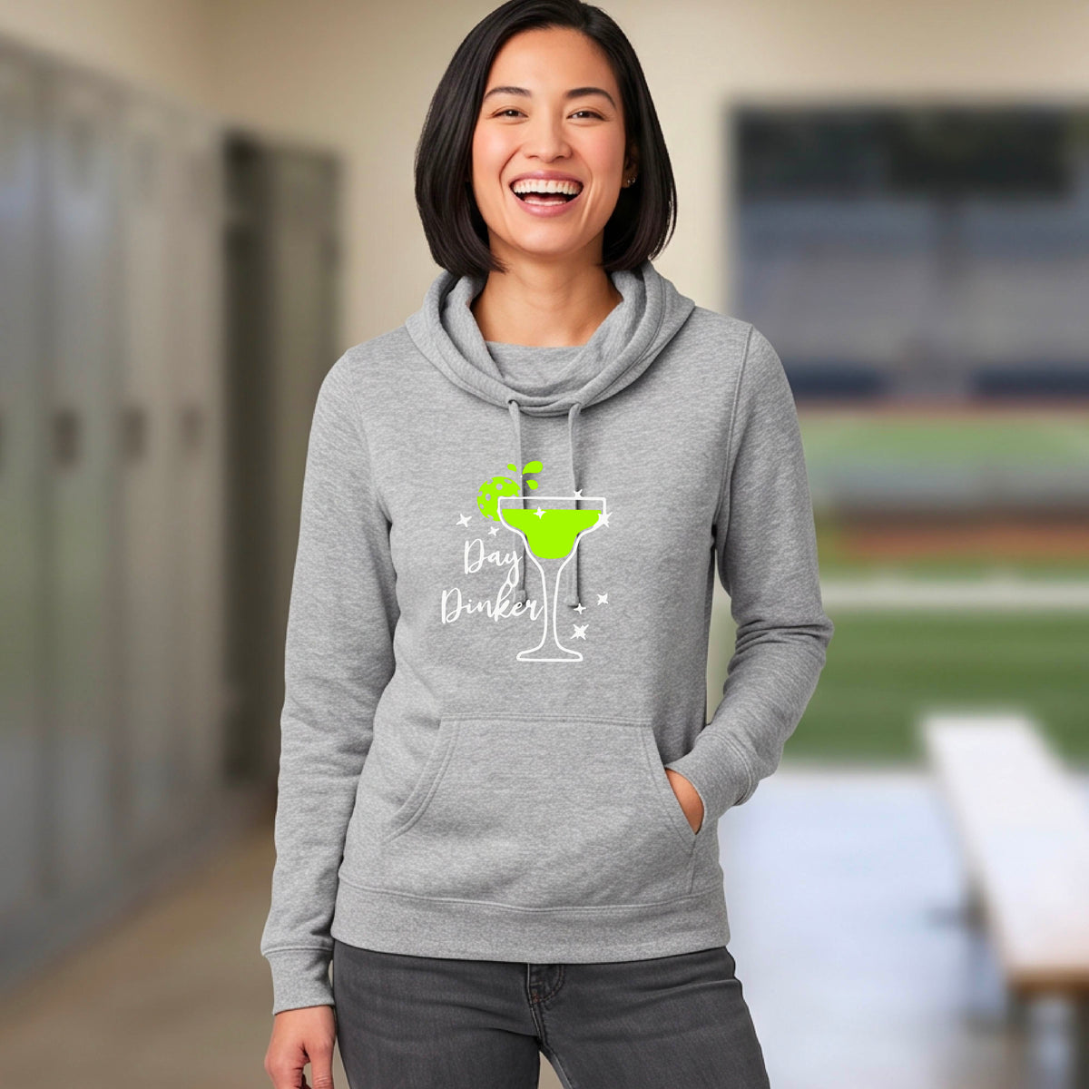 A woman laughing on a pickleball court wearing a gray hoodie with a green cocktail glass graphic and green pickleball lime garnish and the words 'Day Dinker' printed on the front.