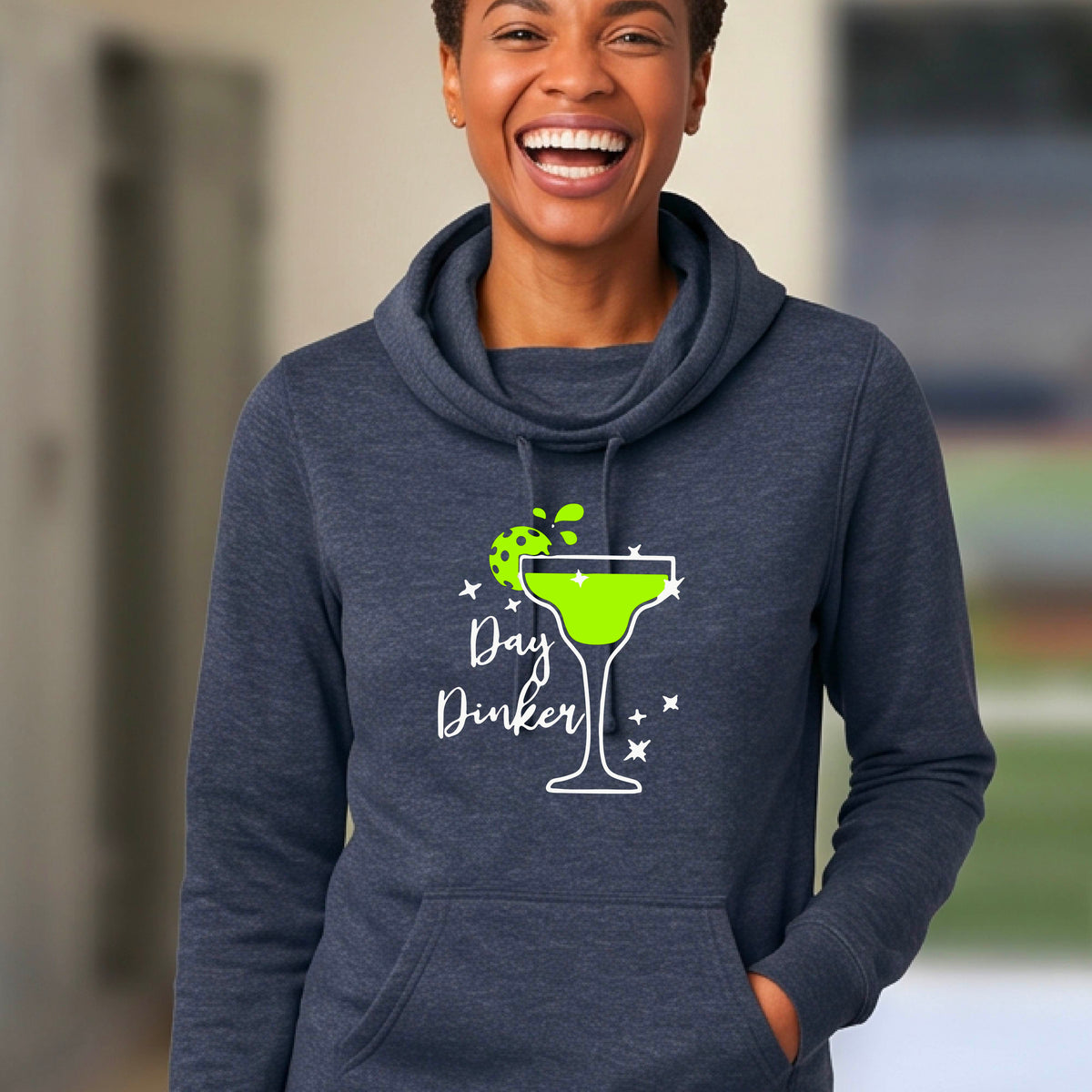 A woman laughing on a pickleball court wearing a navy blue hoodie with a green cocktail glass graphic and green pickleball lime garnish and the words 'Day Dinker' printed on the front.