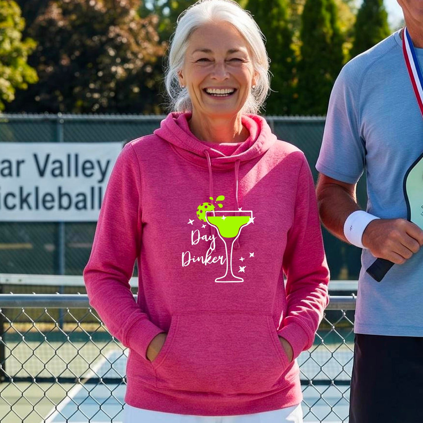 A woman laughing on a pickleball court wearing a pink hoodie with a green cocktail glass graphic and green pickleball lime garnish and the words 'Day Dinker' printed on the front.