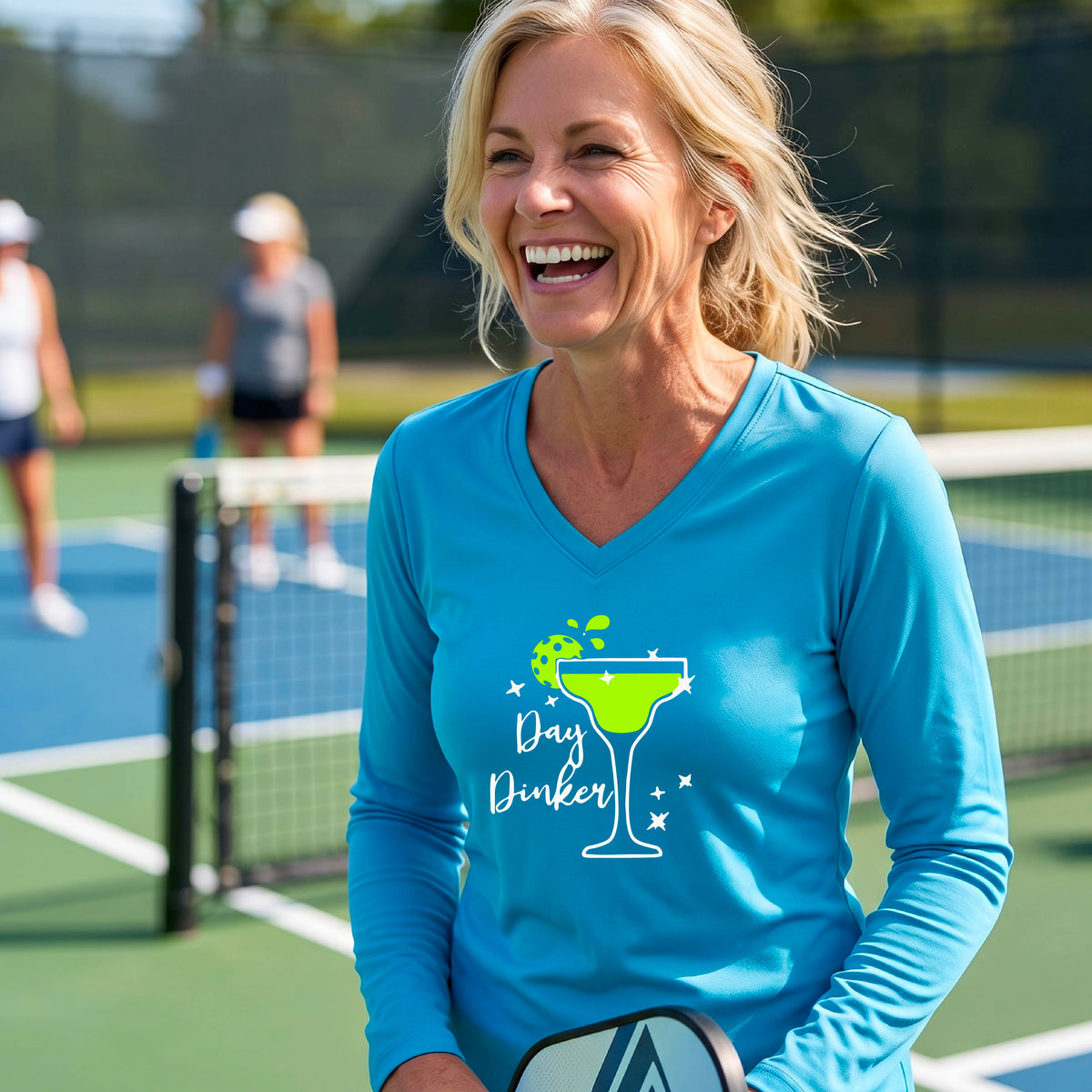 A woman wearing a bright blue long-sleeve V-neck t-shirt with a 'Day Dinker' martini glass graphic in green and white with a pickleball lime garnish. 