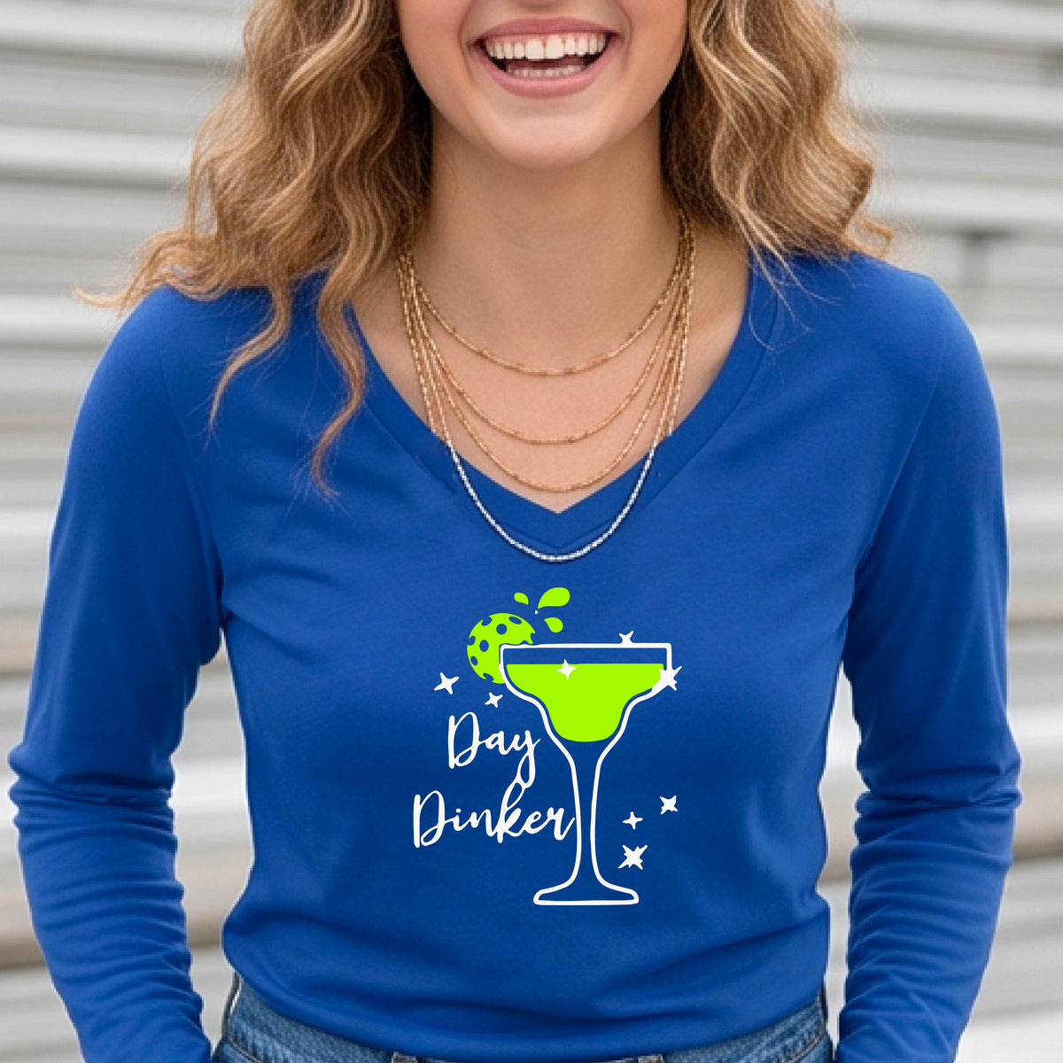 A woman wearing a royal blue long-sleeve V-neck t-shirt with a 'Day Dinker' martini glass graphic in green and white with a pickleball lime garnish. 