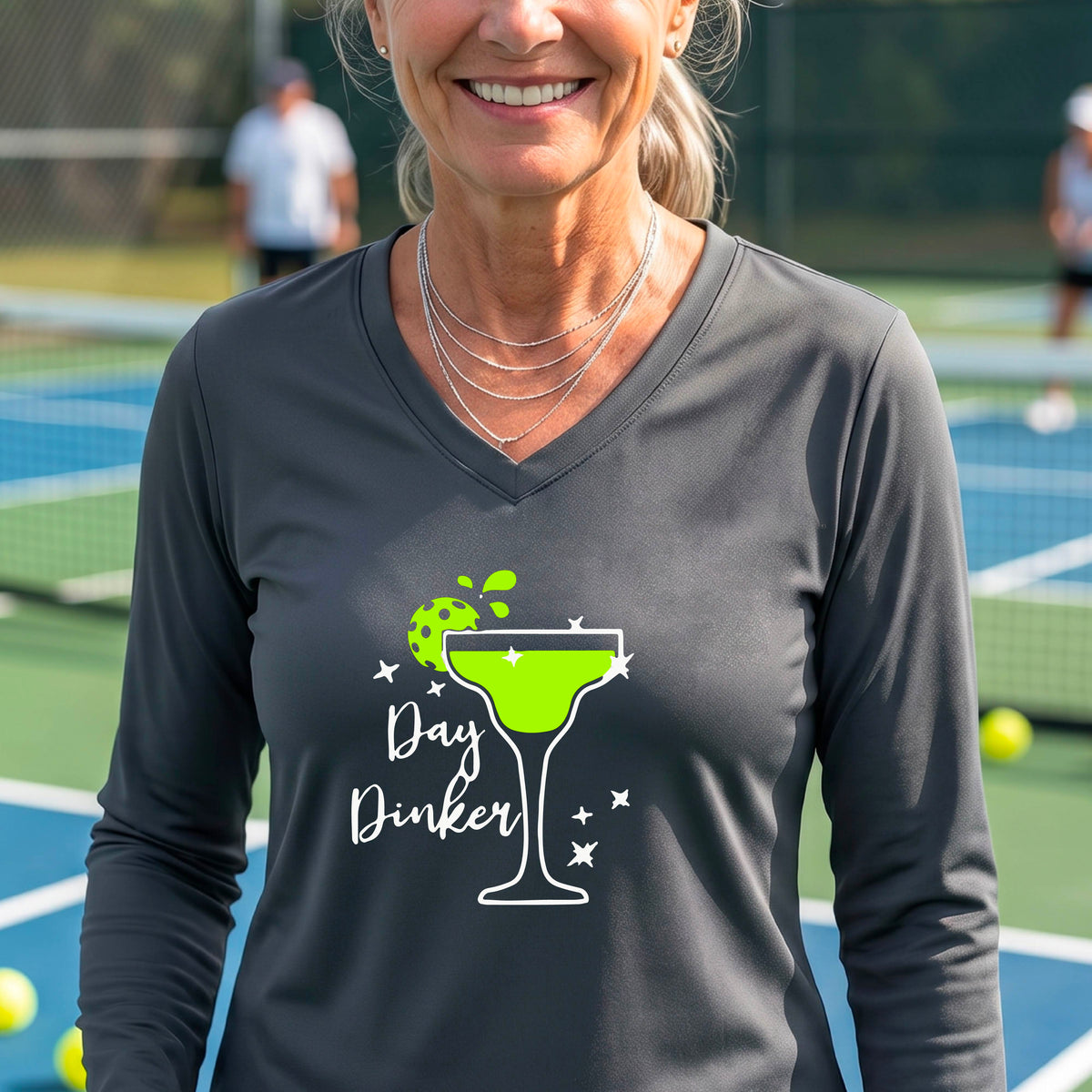 A woman wearing a dark gray long-sleeve V-neck t-shirt with a 'Day Dinker' martini glass graphic in green and white with a pickleball lime garnish. 